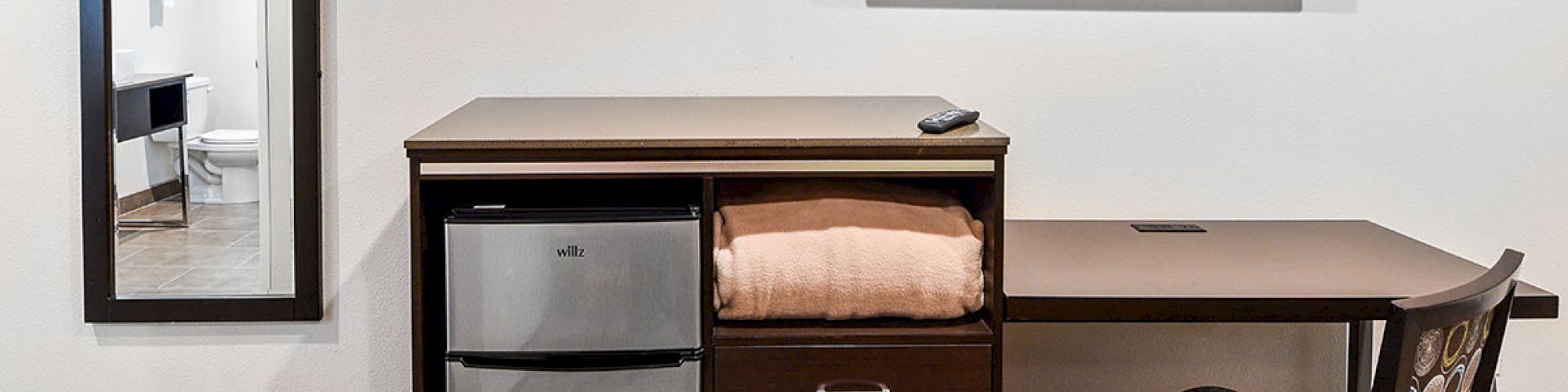 This image shows a hotel room corner with a wall-mounted TV, a floor mirror, a mini-fridge, drawers, and a desk with a chair.