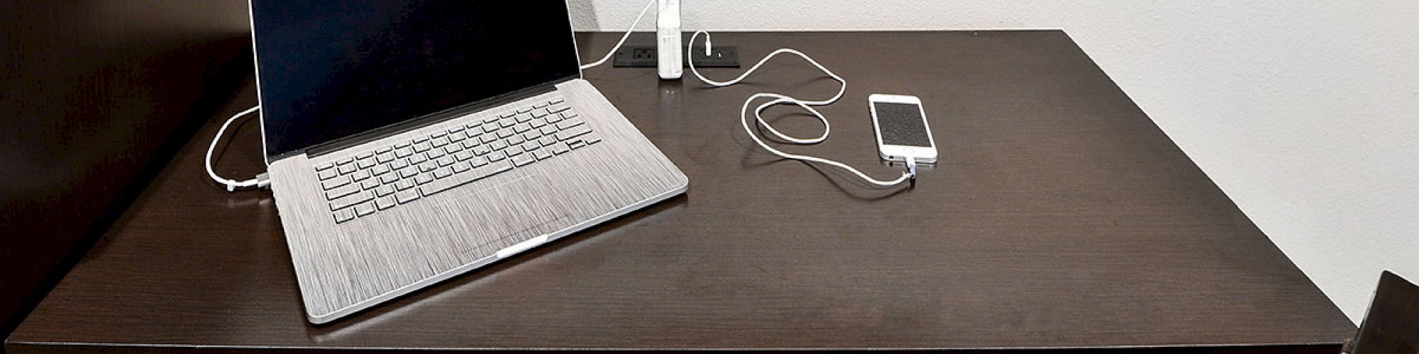 A laptop, smartphone, and charger are on a dark wooden desk with a chair and a cushion with a circular pattern underneath.