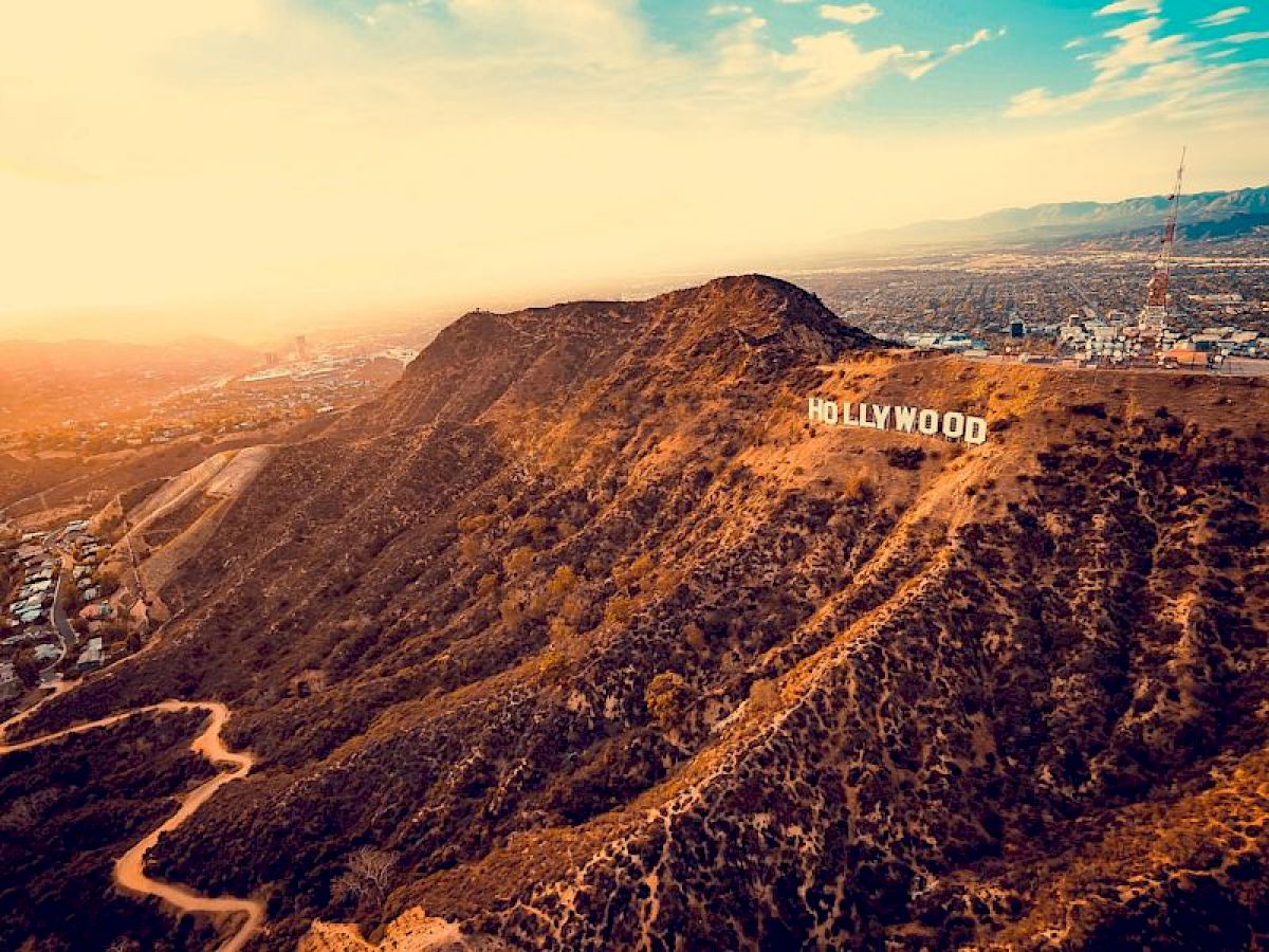 A panoramic view of the Hollywood Hills with the iconic white Hollywood sign, overlooking Los Angeles during sunrise or sunset with a pathway leading up.