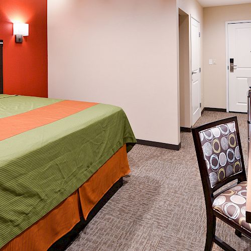 A hotel room features a bed with green and orange bedding, a work desk with a patterned chair, and a door in the background.