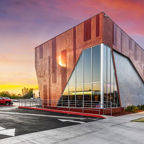 The image shows a modern building with large glass windows and a distinctive angular design, situated by a road during a colorful sunset.