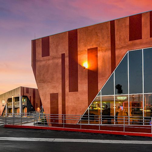 A modern, angular building with geometric patterns and large windows, set against a colorful sunset sky.