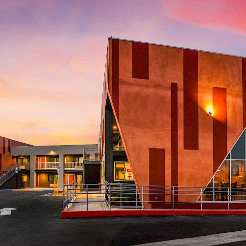 A modern building with a unique geometric design and vibrant colors, surrounded by a parking lot, under a colorful sky at sunset.