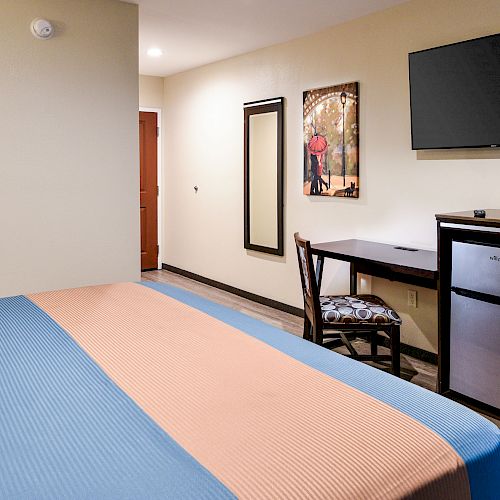 A hotel room features a bed, wall-mounted TV, desk with a chair, mini-fridge, wall mirror, and a framed picture on the wall above the desk.