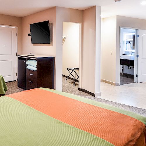 A hotel room with two beds, TV, small table with chairs, open bathroom door, and luggage rack. The beds have green and orange bedspreads.