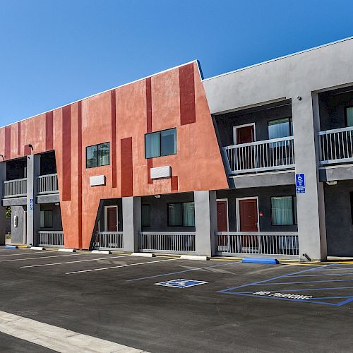 The image shows a modern two-story building with orange and white facades, balconies, and an adjacent parking lot with accessible parking spaces, in clear weather.