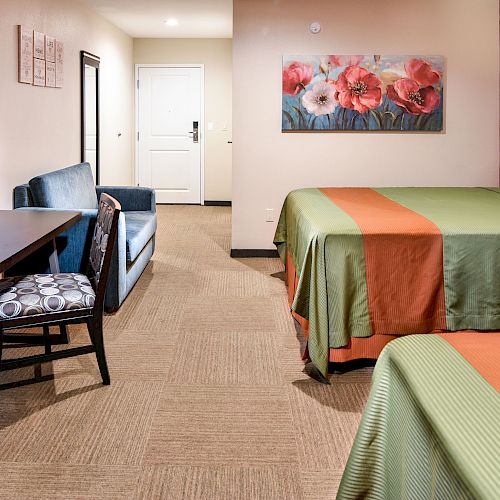 The image shows a hotel room with two beds, a TV, drawers, a sofa, a desk and chair, a floral painting above the beds, and a door in the background.