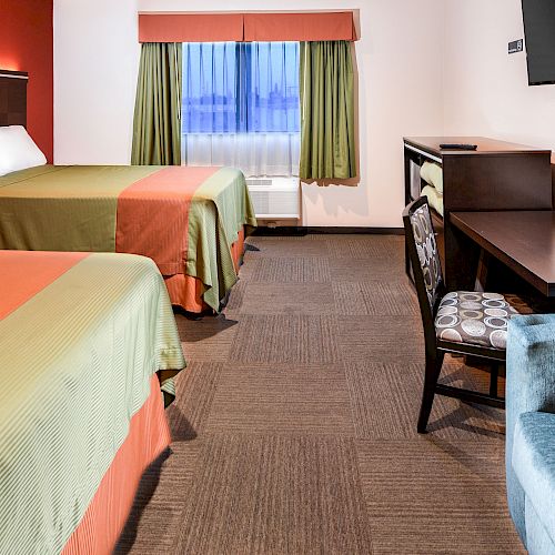 A hotel room with two beds, a window, a desk with a chair, a wall-mounted TV, and an armchair in the foreground.