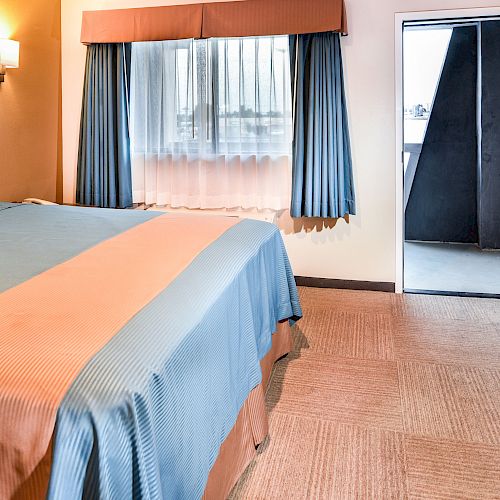 A hotel room with a large bed, bedside lighting, a window with curtains, a partially open door, a mini-fridge, and a nightstand.