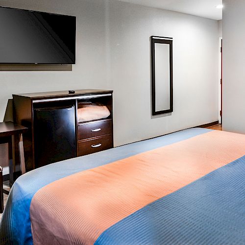 A hotel room with a bed, desk and chair, wall-mounted TV, dresser, and a mirror on the wall, creating a neat and comfortable atmosphere.