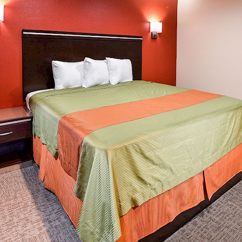 A neatly made bed with a green and orange bedspread, three pillows, and a nightstand against a red wall with two wall-mounted lights.