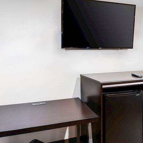 The image shows a simple room setup with a mounted flat-screen TV, a small desk and chair, a cabinet with drawers, and a mini-fridge.