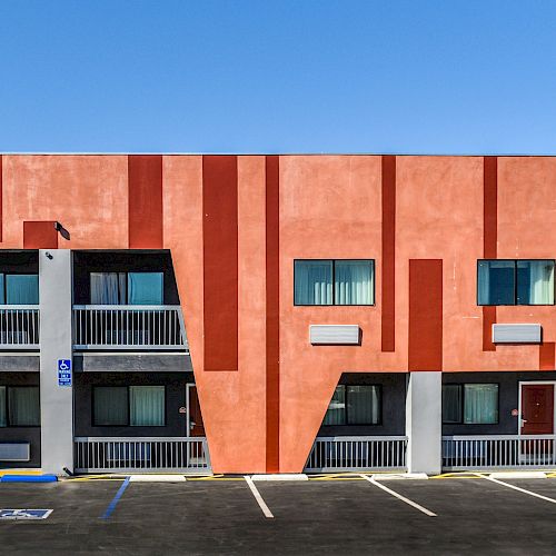 The image shows a two-story building with a unique red and orange geometric design, featuring multiple doors and windows, and an empty parking lot.