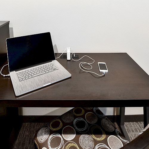 A laptop, phone, and charger cables are on a dark wooden desk with an office chair and a cushion underneath with circular patterns ending the sentence.