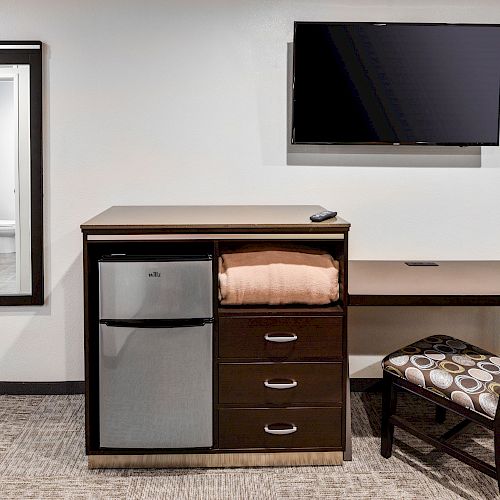 The image shows a wall mirror, a dresser with a mini fridge, drawers, and a folded blanket beside a desk with a chair, and a mounted TV.