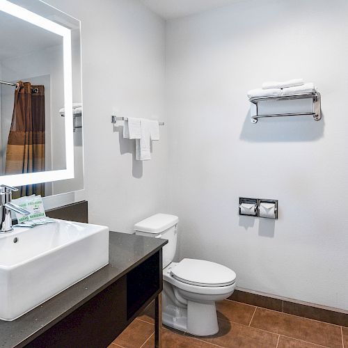 A modern bathroom with a sink, mirror, toilet, towel rack with towels, and a shower with a curtain in the background, ending the sentence.