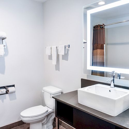 A modern, clean bathroom with a large illuminated mirror, towel racks, a toilet, a sink with a vanity, and a shower curtain.