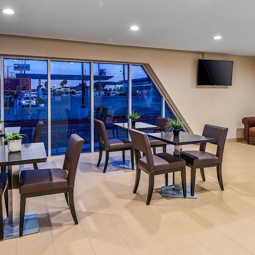 The image shows a modern waiting area with tables, chairs, a brown couch, decorative wall art, and large windows offering a view outside.