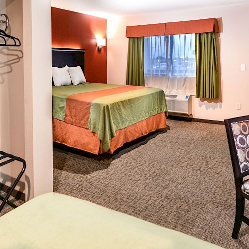 A hotel room with a bed, chair, table, ironing board, and closet area. The window has green and orange curtains, and the walls are beige and red.