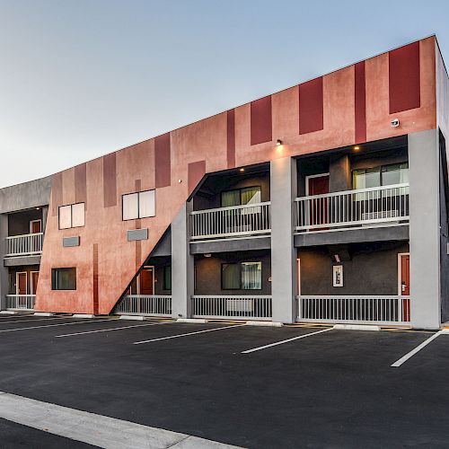 The image shows a modern two-story building with a colorful exterior, balconies, and an adjacent parking lot with marked spaces.