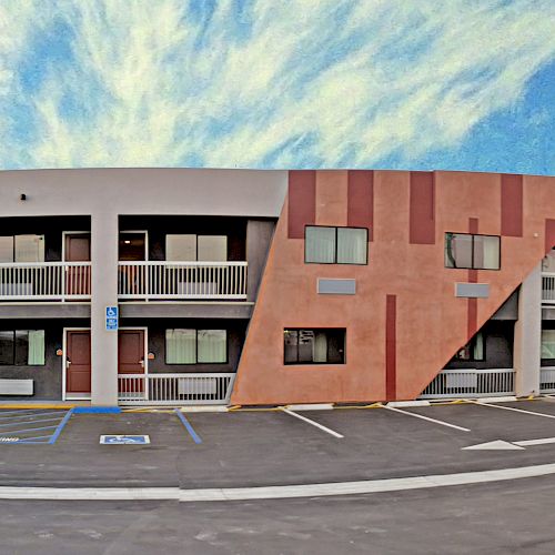 The image features a two-story motel with a modern facade, a parking lot with several cars, a cloudy blue sky, and accessible parking spaces.