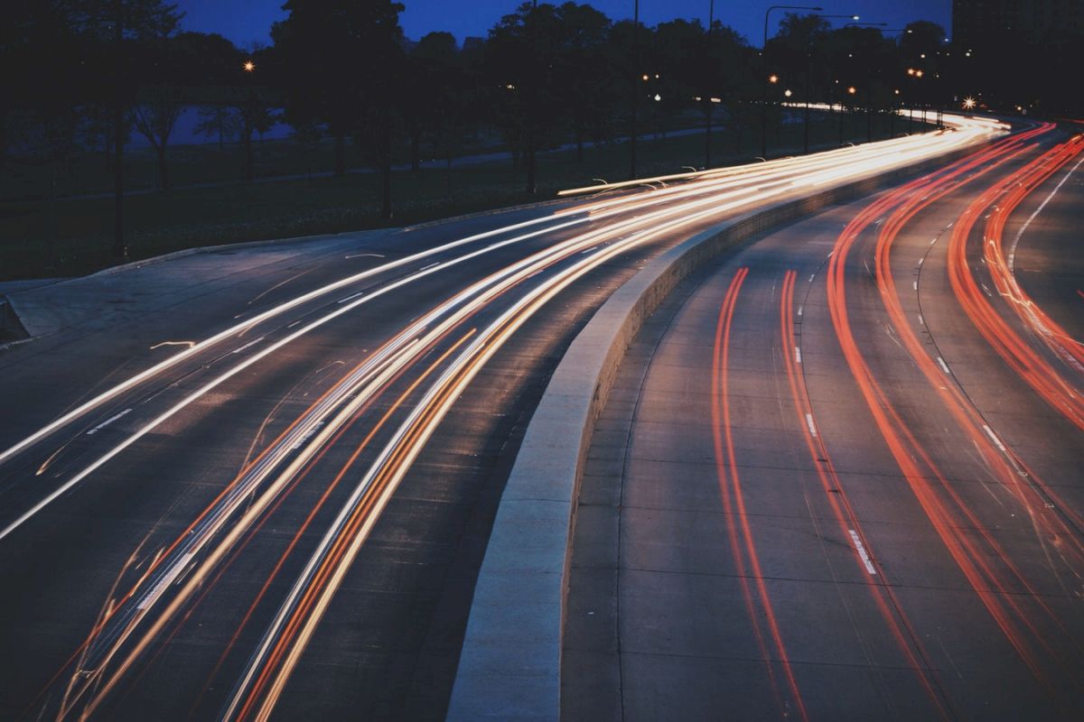 A nighttime long-exposure photo of a road shows light trails from vehicles, creating bright streaks against the dark surroundings.