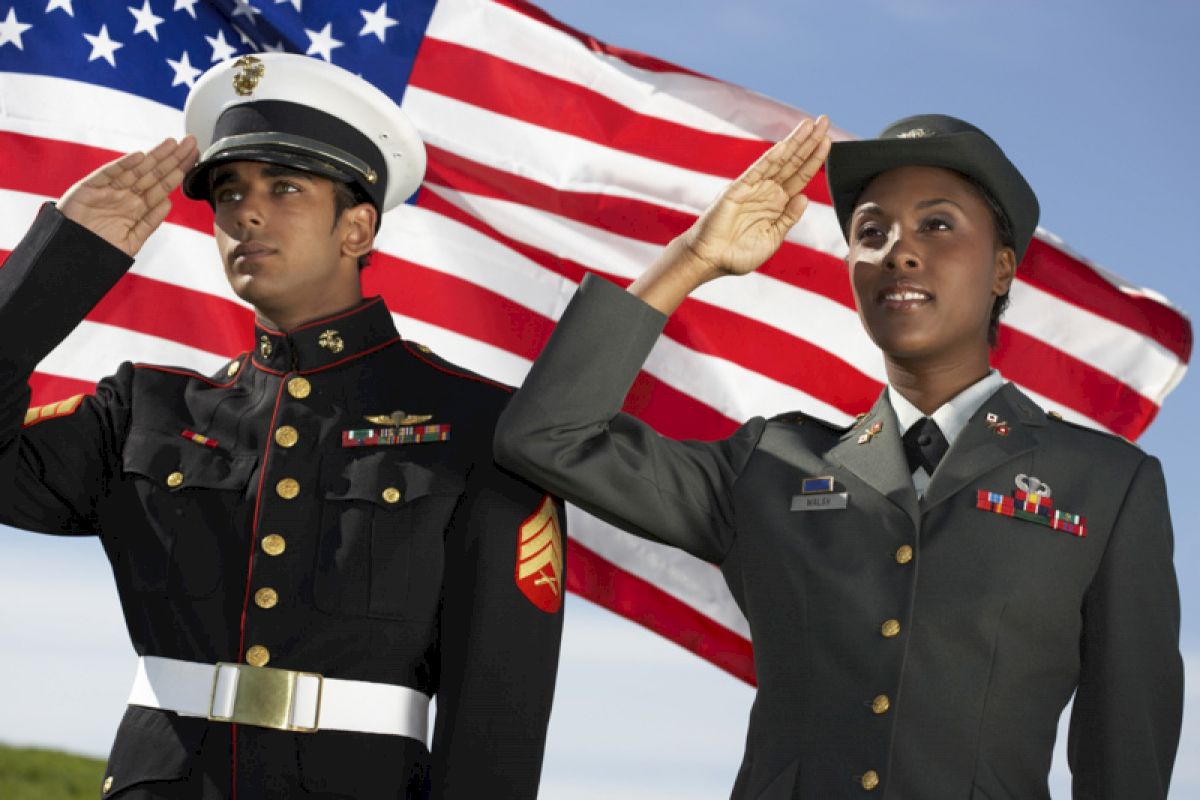 Two military personnel are saluting, standing in front of an American flag on a sunny day.