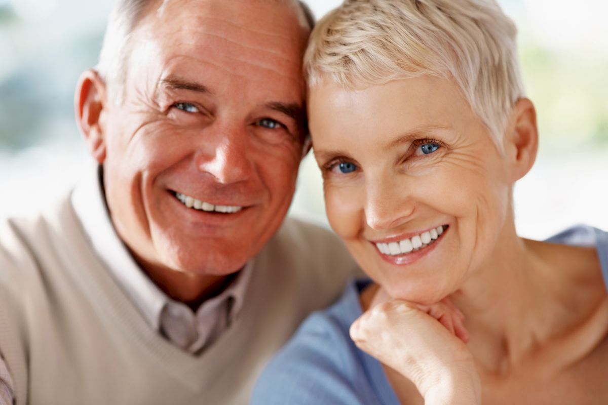 This image shows an elderly couple, both smiling and looking at the camera, sitting closely together in a warm and positive setting.