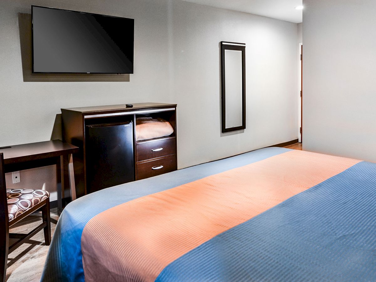 A hotel room features a bed with a blue and orange cover, a wall-mounted TV, a desk with a chair, a dresser, and a mirror on the wall.