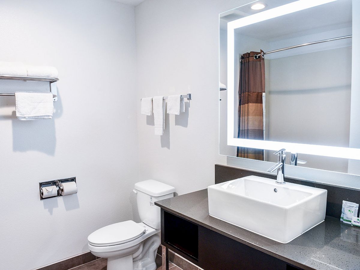 A clean, modern bathroom with a sink, illuminated mirror, toilet, towel rack, and a shower with a curtain seen in the background.