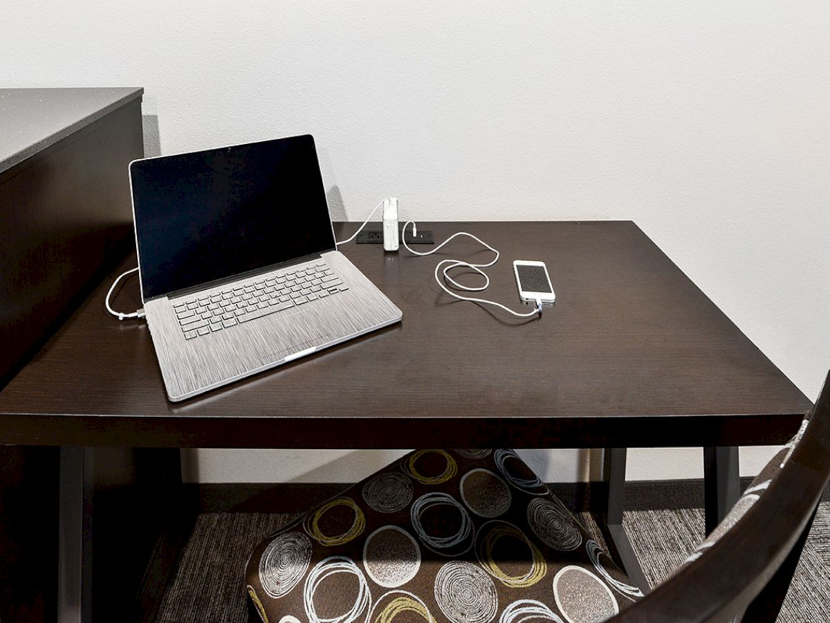 A dark wooden desk with a laptop, an upright phone charger holding a smartphone, and a patterned cushion underneath the desk.