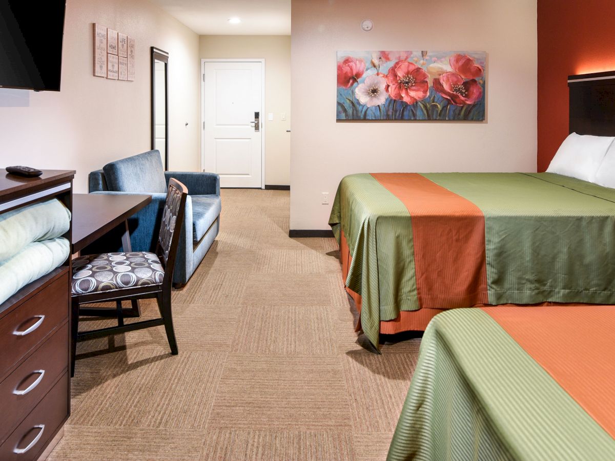 A hotel room with two beds, colorful bedspreads, a TV, a chair, a desk, and a bathroom in the background, and a floral painting on the wall.