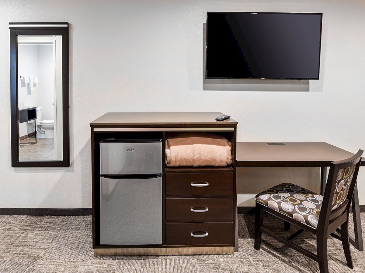 A hotel room setup with a mini-fridge, drawers, a desk, chair, wall-mounted TV, and a mirror, with a bathroom visible in the mirror reflection.