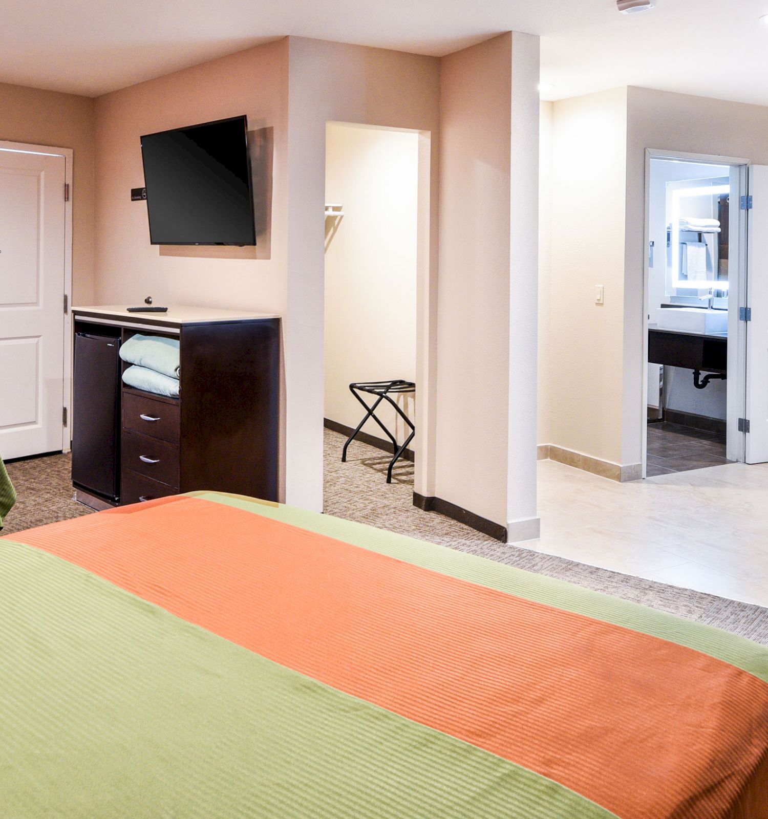 The image shows a hotel room with two beds, a wall-mounted TV, an open closet, a table with chairs, and a view into the bathroom area.