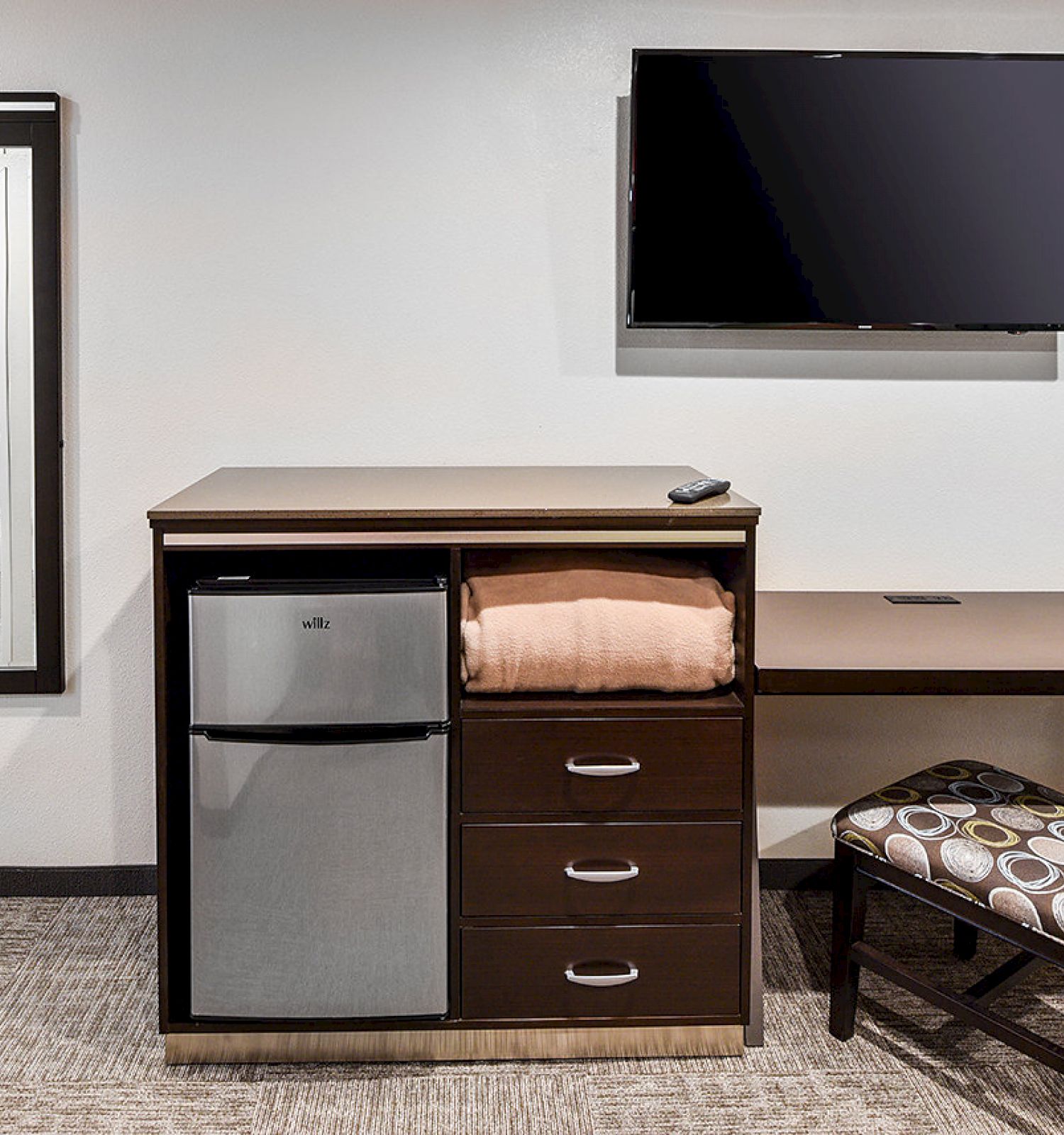 The image shows a room with a desk, a chair, a small fridge, a TV on the wall, and a mirror reflecting a glimpse of a doorway.