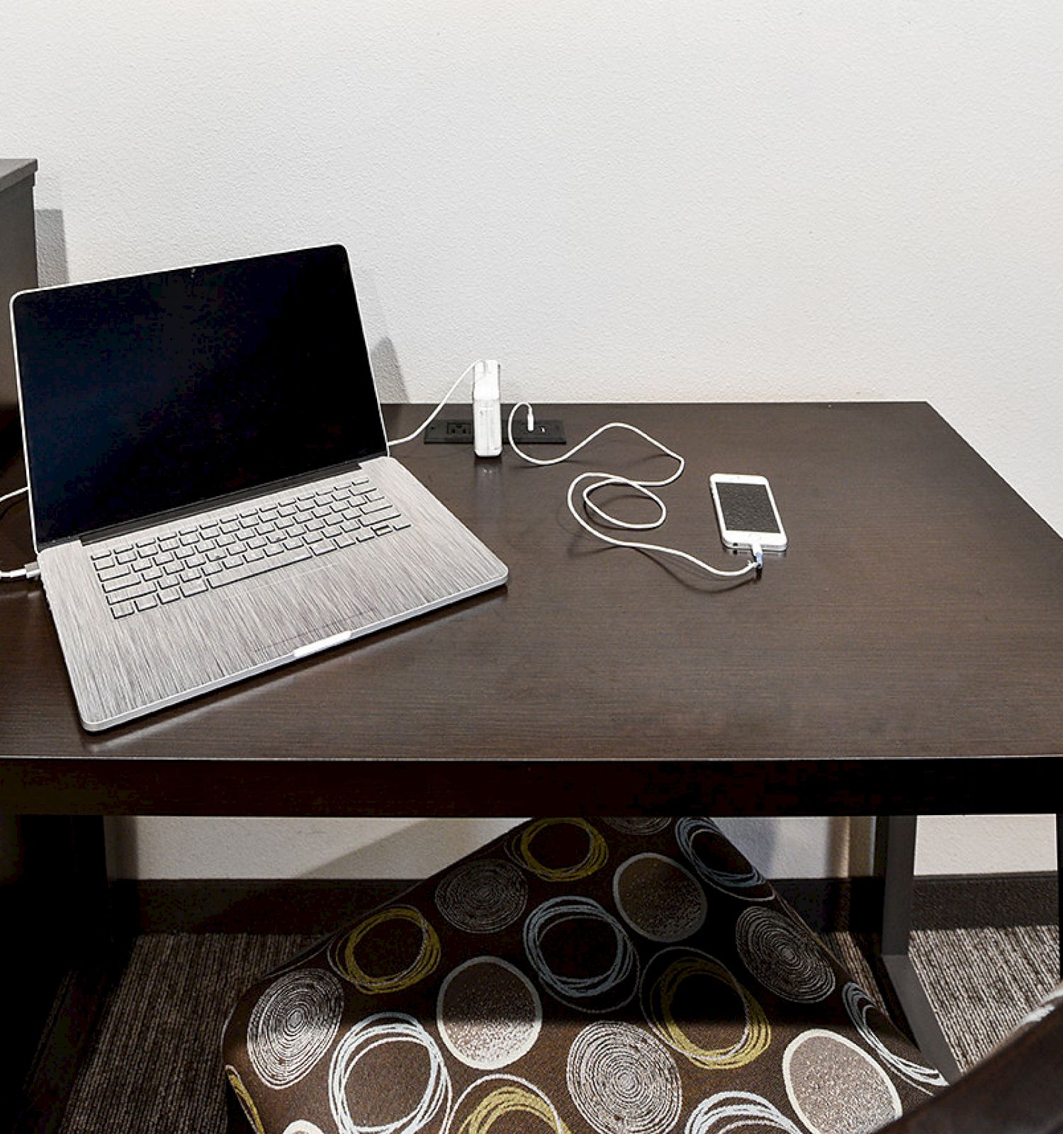 A laptop, smartphone, wireless earphones, and a charging station are on a dark brown desk. The setup includes a patterned pillow under the table.