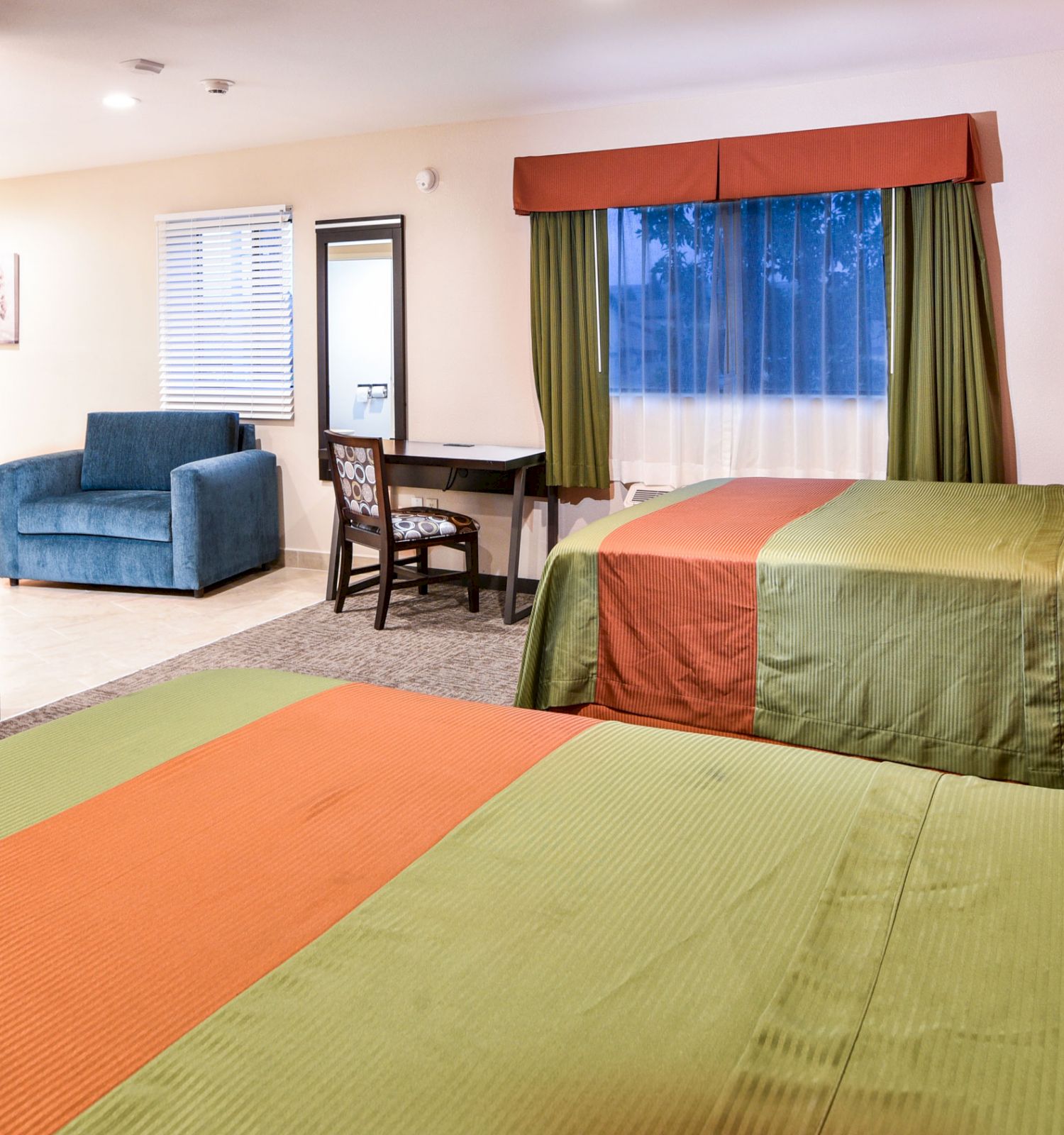 A hotel room with two beds, a TV, a dresser, a table and chairs, a sofa, and a window with green curtains. The room has a modern, clean look.