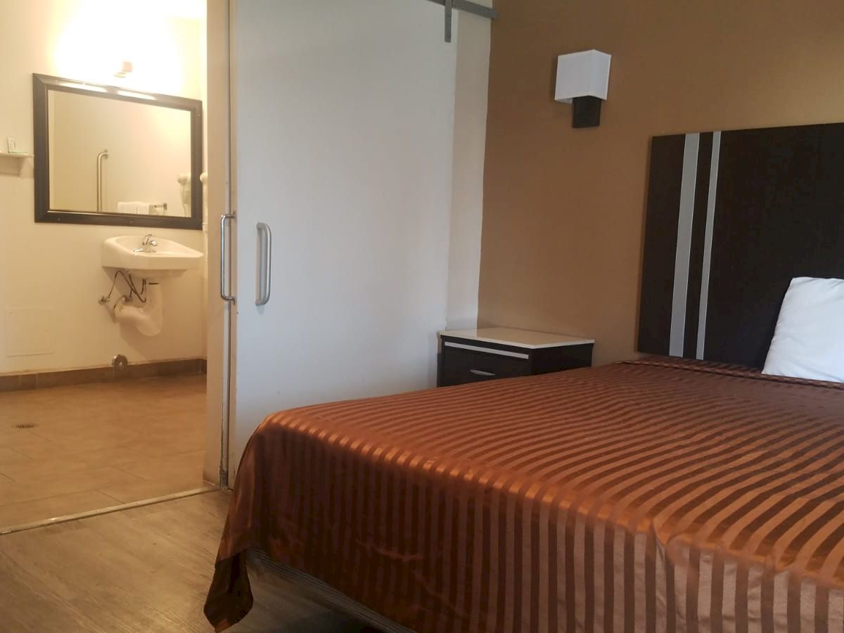 The image shows a hotel room with a striped bedspread, a nightstand, and an open door leading to a bathroom with a sink and mirror, light on.