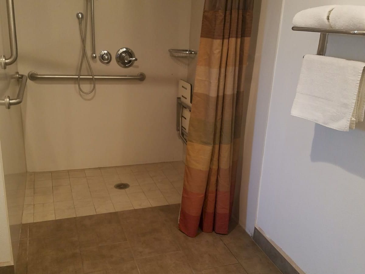 This image shows an accessible bathroom with a walk-in shower equipped with grab bars, a shower chair, and a curtain. Towels are on a rack.