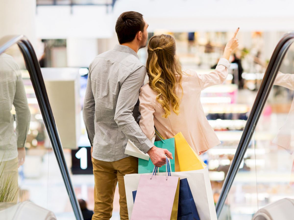 A couple is riding an escalator in a shopping mall, holding shopping bags and looking at something ahead, while the woman points forward.