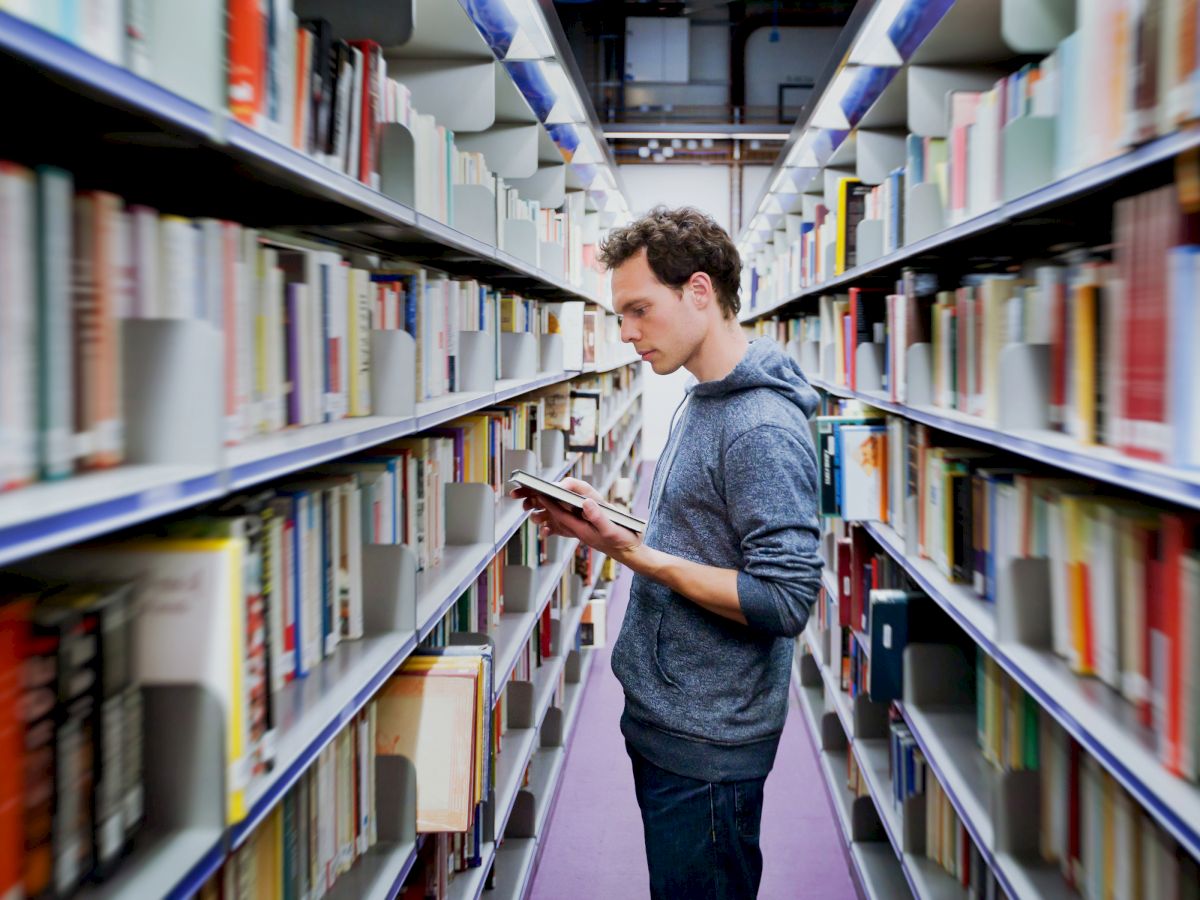 A person is standing between bookshelves in a library, reading a book. He is surrounded by rows of neatly arranged books on both sides.