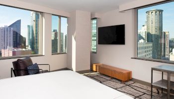A modern hotel room with large windows showing a cityscape, featuring a bed, an LCD TV, chair, and a cabinet against the wall ending the sentence.