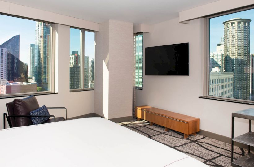 A modern hotel room with large windows showing a cityscape, featuring a bed, an LCD TV, chair, and a cabinet against the wall ending the sentence.