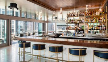 A modern bar with a wooden counter and blue barstools, well-stocked with various bottles of alcohol, large windows, and cityscape views.