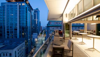 A modern cityscape at dusk features tall buildings and a rooftop terrace with seating and tables overlooking a bustling street.