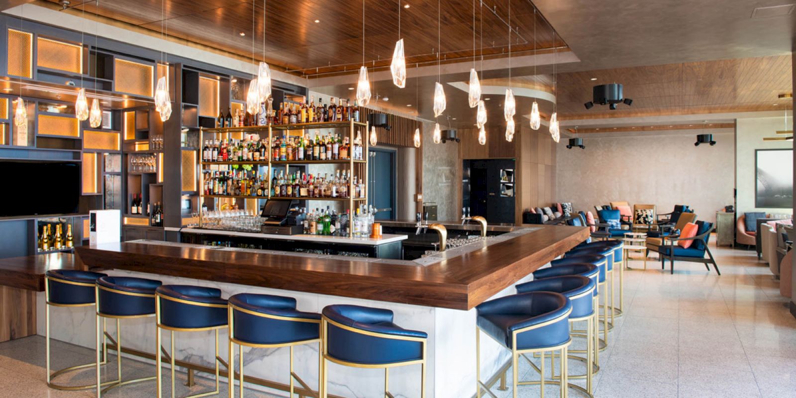 A modern bar with high chairs, hanging lights, shelves of liquor bottles, and a seating area in the background ending the sentence.