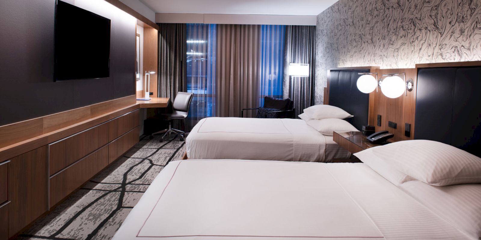 The image shows a modern hotel room with two double beds, a large TV, a desk with a chair, and a patterned wall.