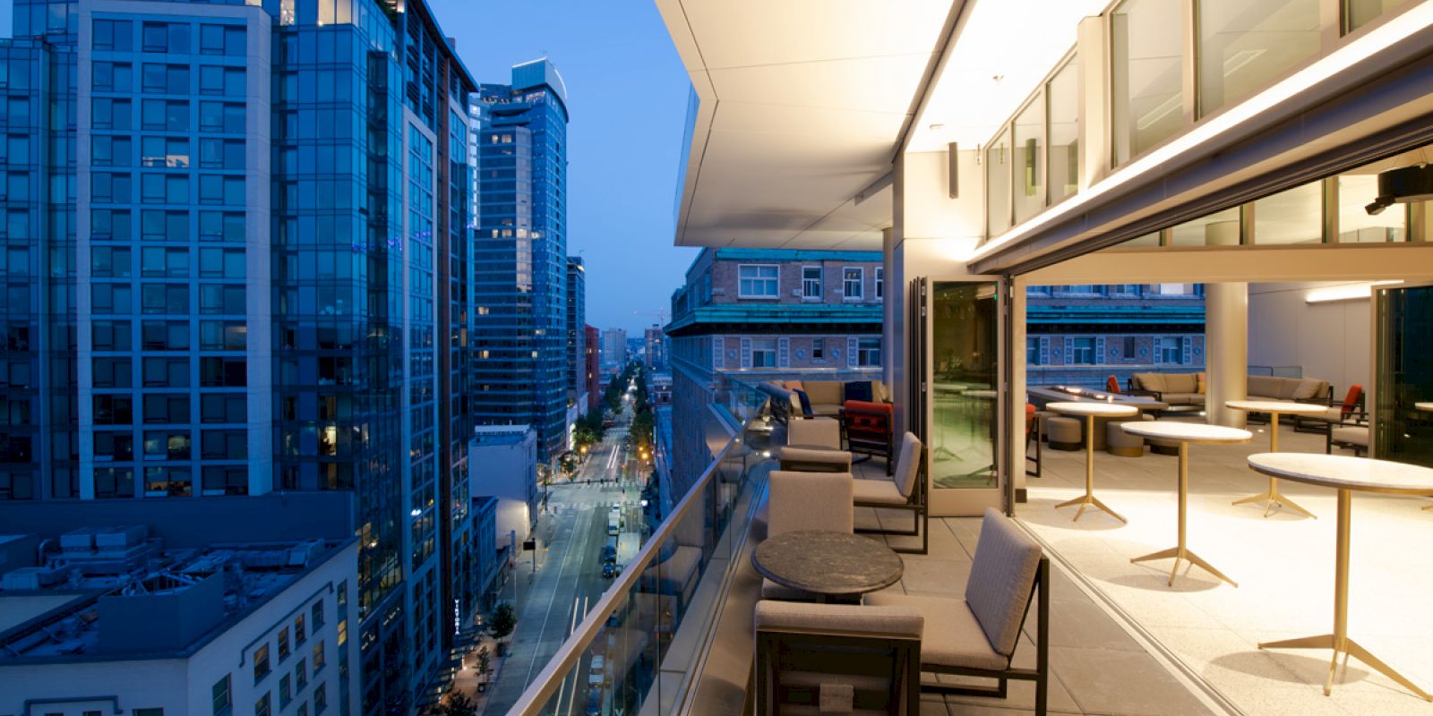 A modern urban balcony overlooks a cityscape with high-rise buildings, streetlights, and minimalistic furniture, creating a sleek evening ambiance.