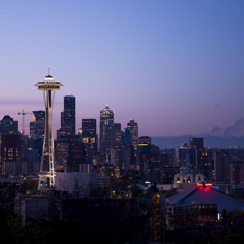 A panoramic view of Seattle's skyline at dusk, featuring the Space Needle prominently, with Mount Rainier visible in the background and city lights aglow.