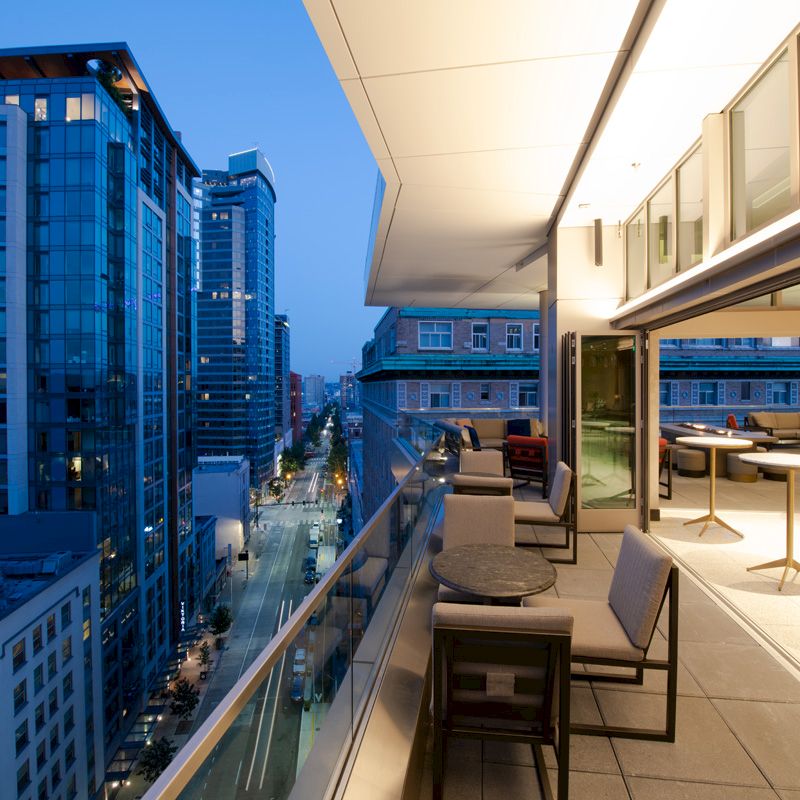 A modern cityscape at dusk features a high-rise building balcony with tables and chairs, overlooking a street lined with tall buildings.